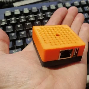 Drobné oranžové zařízení s ethernetovým portem položené na dlani před klávesnicí