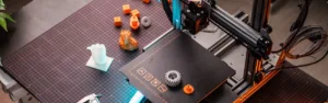 Otevřená tiskárna Prusa 3D tiskne oranžowe prvky vedle hotových výtisků