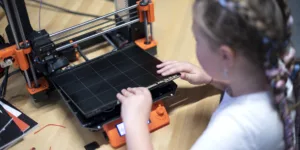 Dítě obsluhuje oranžovočernou 3D tiskárnu Prusa u pracovního stolu