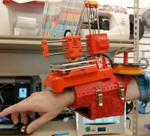 Kompaktní oranžová 3D tiskárna umístěná kolem předloktí člověka
